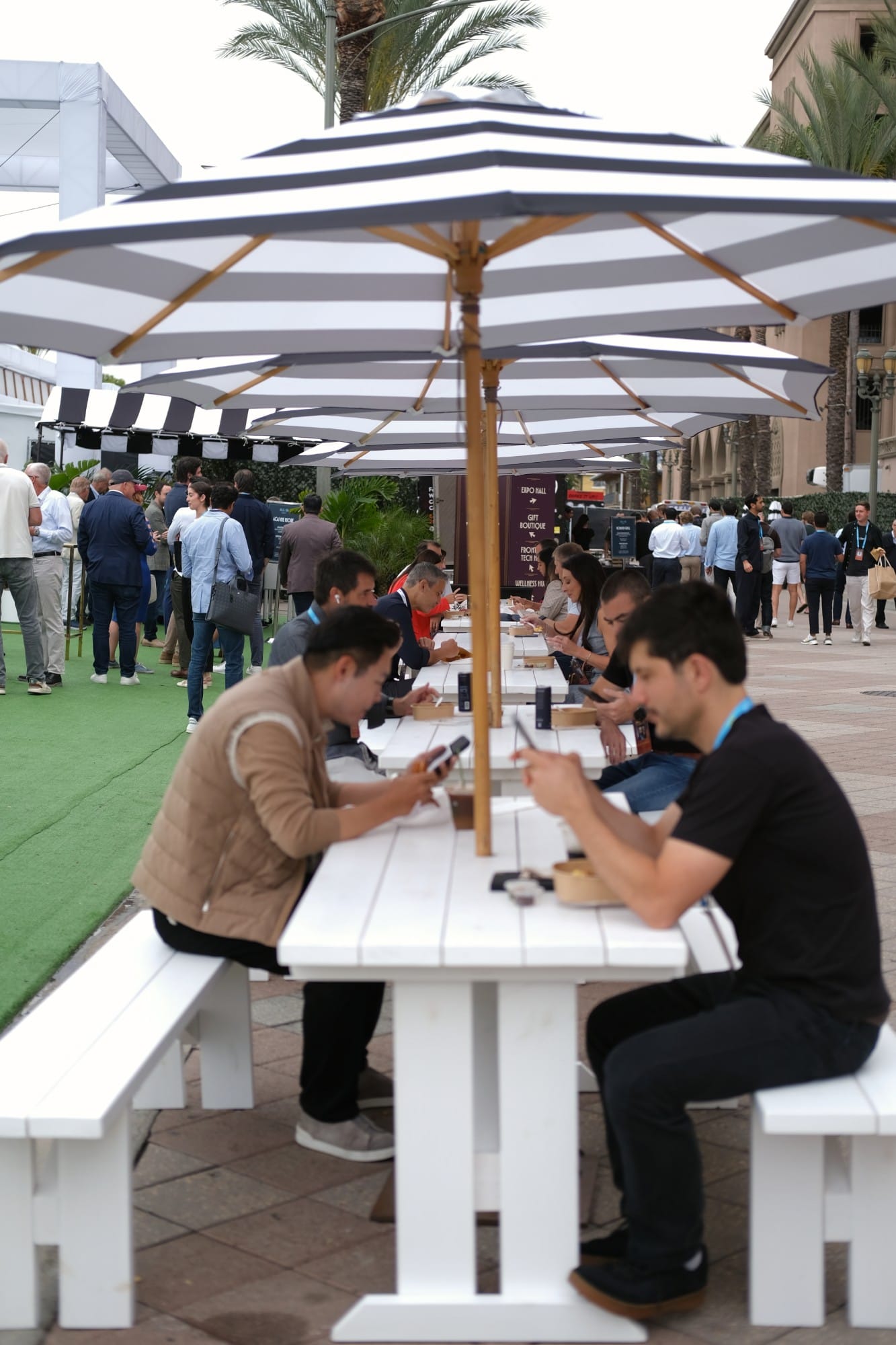 Attendees at long lunch tables under striped umbrellas, each looking down at their own phone